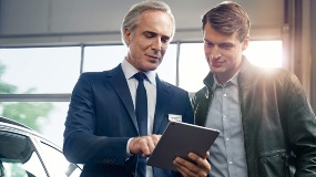 Zwei Männer betrachten ein Tablet in einer Autowerkstatt, links ein Auto im Hintergrund.