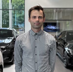 Mann in kariertem Hemd steht in einem Autohaus, im Hintergrund sind Autos und ein Bildschirm.