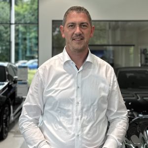 Mann in weißem Hemd in einem Autohaus mit Autos im Hintergrund.