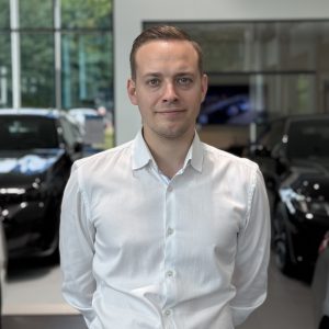 Ein Mann in weißem Hemd steht in einem Autohaus mit schwarzen Autos im Hintergrund.