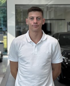 Ein Mann in weißem Polohemd steht in einem Autohaus, im Hintergrund sind Autos zu sehen.