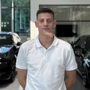 Ein Mann in weißem Polohemd steht in einem Autohaus mit BMW-Fahrzeugen im Hintergrund.