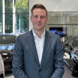 Ein Mann im Anzug steht in einem modernen Autohaus, im Hintergrund sind BMW-Fahrzeuge.