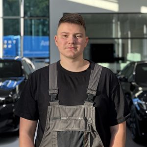 Ein Mann in Arbeitskleidung steht in einem BMW Autohaus mit Autos im Hintergrund.