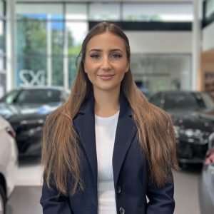 Frau in blauem Blazer steht in einem BMW-Autohaus; Autos im Hintergrund.