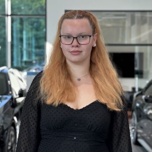 Frau mit langen Haaren und Brille in einem Autohaus neben BMW-Fahrzeugen.