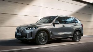 Silberner BMW X5 SUV fährt dynamisch vor einer grauen Wand.