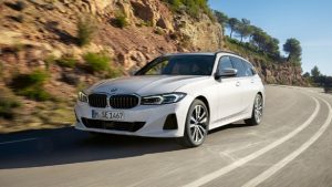 Weißer BMW auf geschwungener Landstraße bei Sonnenschein mit Bäumen und Felsen im Hintergrund.