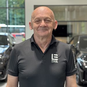 Mann in schwarzem Polohemd mit Logo in einem Autohaus mit unscharfen BMW-Fahrzeugen im Hintergrund.