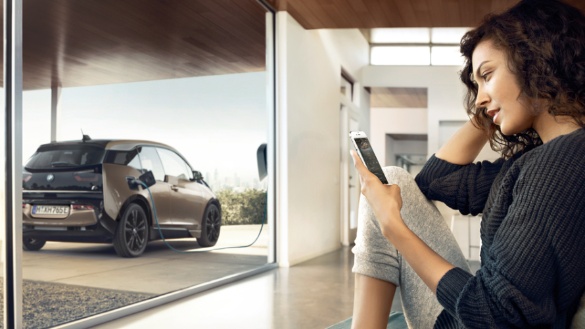 Eine Frau sitzt drinnen, schaut auf ihr Smartphone; draußen lädt ein BMW i3 an einer Ladestation.