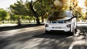 Weißer BMW i3 auf einer Straße, umgeben von Bäumen bei Sonnenlicht.