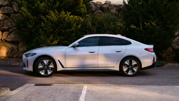 Weißer BMW i4 steht seitlich auf einer Straße, umgeben von Bäumen und einer Steinmauer.