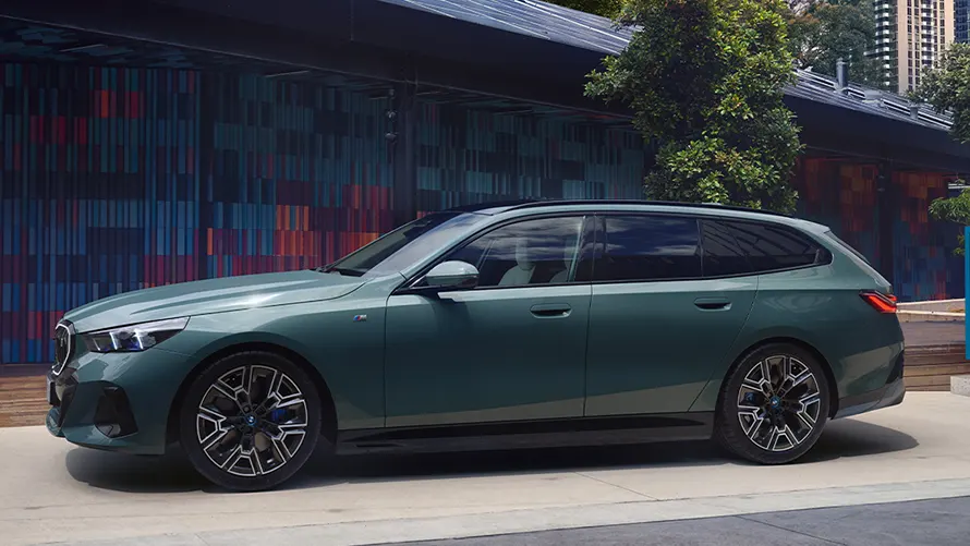 Grüner BMW Kombi, seitlich, vor modernem Gebäude mit bunten Wänden und Bäumen.