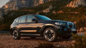 Ein schwarzer BMW iX3 steht vor einer Berglandschaft bei Sonnenuntergang auf einem Kiesweg.