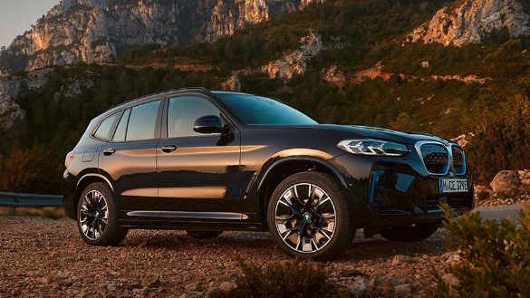 Ein schwarzer BMW iX3 steht vor einer Berglandschaft bei Sonnenuntergang auf einem Kiesweg.