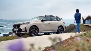 Ein beiger BMW X3 steht auf einer Küstenstraße, im Hintergrund das Meer und eine Frau.