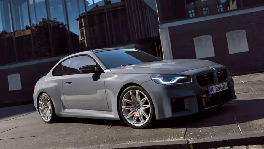 Graues BMW 2er M Coupé auf einer Straße, modernes Gebäude im Hintergrund.