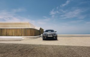 Grauer BMW i4 auf hellem Parkplatz vor modernem Gebäude, blauer Himmel im Hintergrund.