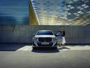 Silberner BMW iX1 vor moderner Architektur, Frau öffnet Fahrertür.