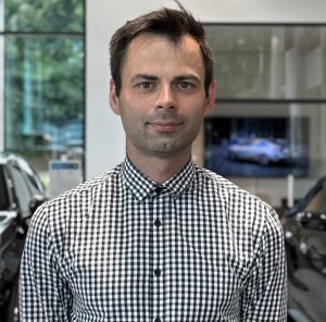 Ein Mann in kariertem Hemd steht in einem Autohaus mit unscharfen Autos im Hintergrund.