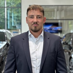 Mann in Anzug steht in einem Ausstellungsraum mit Autos im Hintergrund.