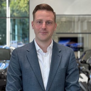 Mann in grauem Anzug steht in einem Autohaus vor unscharfem Hintergrund mit Autos.