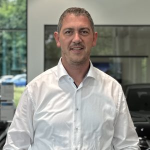 Ein Mann in weißem Hemd steht lächelnd in einem Autohaus mit Glasfenstern im Hintergrund.